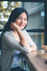 Beautiful attractive young Asian woman holding a cup of coffee in hand and sitting on sofa at cafe in the morning, vintage color tone. 