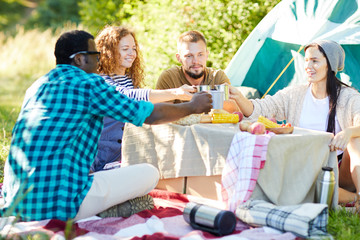 Young friends clinking with metal mugs with hot tea after lunch on their backpack trip