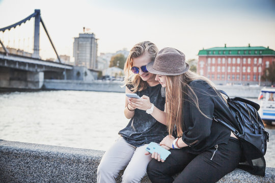 Two young stylish girls female teenagers 18 years old on the background of a European city and a river with a smartphone in their hands, communication and navigation application,
