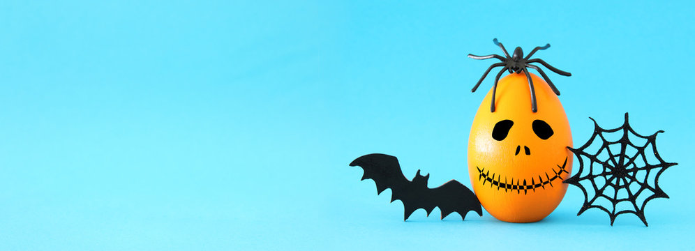 minimal and funny Halloween holiday concept. Orange egg with scary cute face, spiderweb, bat and spider on top. Banner.