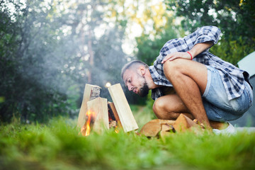 Young active man blowing on fire while squatting by heap of firewoods in camp