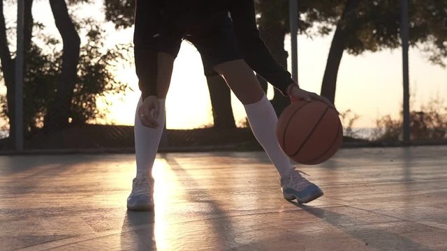 Close up footage of a female athlete legs in white golf socks and sneakers. Female baasketball player bouncing ball from hand to hand., practicing cross legs exercise. Sun shines on the background