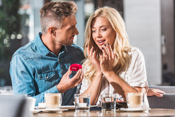 handsome boyfriend proposing shocked girlfriend and showing red ring box in restaurant