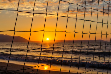 Naklejka premium View of the sunrise through the volleyball net. Early morning , dramatic sunrise over sea and mountain.