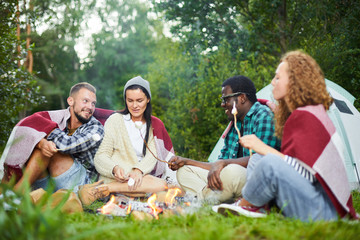 Group of cheerful young backpackers enjoying evening by campfire while having dessert