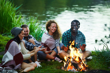 Two young couples of backpackers sitting close to campfire, relaxing and enjoying evening by waterside