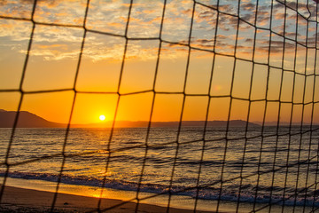 Naklejka premium View of the sunrise through the volleyball net. Early morning , dramatic sunrise over sea and mountain.