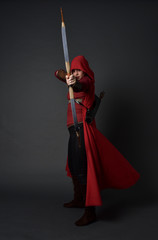 full length portrait of brunette girl wearing red medieval costume and cloak, holding a bow and...