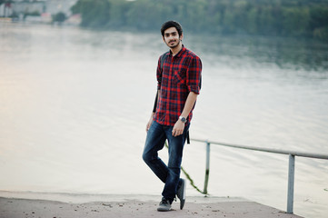 Young indian student man at checkered shirt and jeans with backpack posed on evening city against lake.