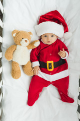 top view of cute little baby in santa suit lying in crib with teddy bear and looking at camera
