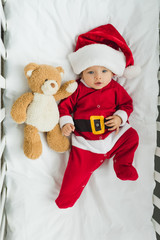top view of beautiful little baby in santa suit lying in crib with teddy bear