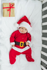 top view of happy little baby in santa suit lying in crib with christmas gift