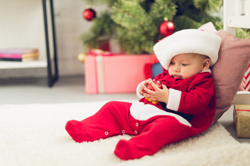 adorable little baby in santa suit sitting on floor with blurred christmas tree on background