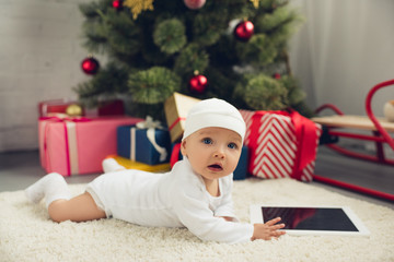 Obraz na płótnie Canvas cute little baby with tablet lying on floor with christmas gifts and tree blurred on background