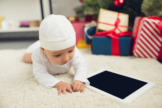 adorable little baby with tablet lying on floor with christmas gifts blurred on background