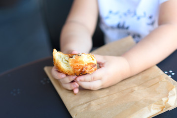 Kid eating croissant.Close up hand Little girl having breakfast in the cafe on the city street. The child is eating a croissant. Breakfast black table in restaurant eco paper bag.