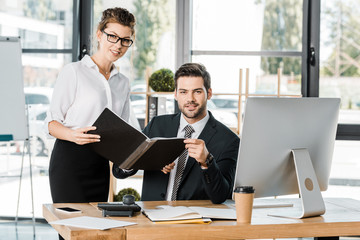businessman sitting at table and looking at camera, businesswoman holding folder with documents in office