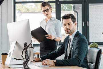 businessman sitting at table and looking at camera, businesswoman holding folder with documents in office