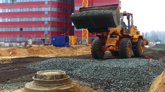 Bulldozer unloading broken stone on ground. Infrastructure construction near new commercial building. Wheel loader excavator working on construction site. Heavy machinery in industrial roadworks