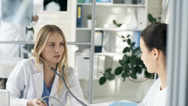 Tilt down shot of young Caucasian female doctor in lab coat smiling and talking to patient while checking blood pressure with sphygmomanometer