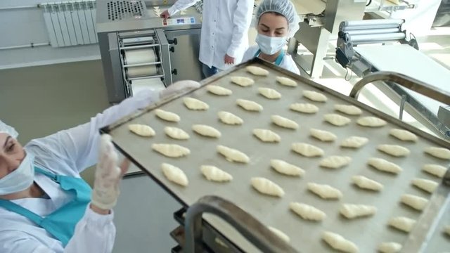High angle view of two female factory workers in protective clothing, aprons, bouffant mob caps, and face masks putting trays with croissants on baking rack