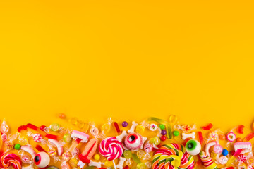 Fototapeta na wymiar Assorted teeth & eyeball shaped candy spread on yellow background, jelly spider, gummy worms, sugar bones, round lollipop and other mixed candy, bloody finger. Top view, copy space, close up, flat lay