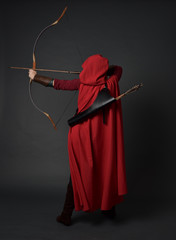 full length portrait of brunette girl wearing red medieval costume and cloak, holding a bow and...