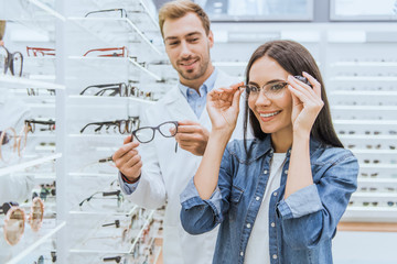 happy woman choosing eyeglasses while male oculist standing near with another eyeglasses in optica - obrazy, fototapety, plakaty
