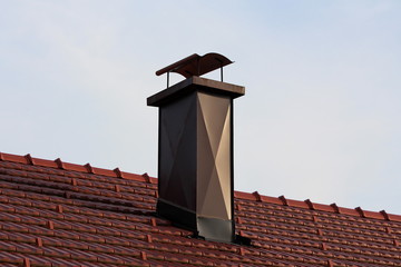 Modern house chimney with brown metal protection on all sides and wavy shield on top surrounded with new red roof tiles at sunset