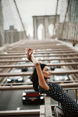 Portrait of gorgeous and tender Indian woman in black dress posing before the Brooklyn bridge...