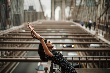 Portrait of gorgeous and tender Indian woman in black dress posing before the Brooklyn bridge somewhere in New York