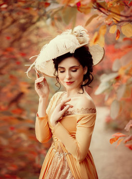 A young brunette woman with an elegant, hairstyle in a hat with a strass feathers. Lady in a yellow vintage dress walks through the autumn landscape. Artistic portrait