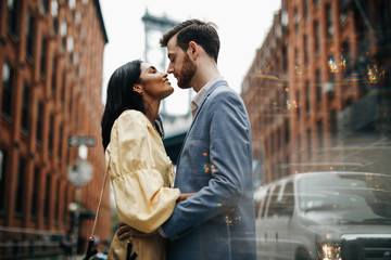 Love story in New York. Gorgeous couple of American man with beard and tender Eastern woman hug each other before the cityscape of Brooklyn bridge somewhere in New York - obrazy, fototapety, plakaty