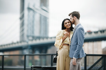 Love story in New York. Gorgeous couple of American man with beard and tender Eastern woman hug each other before the cityscape of Brooklyn bridge somewhere in New York - obrazy, fototapety, plakaty