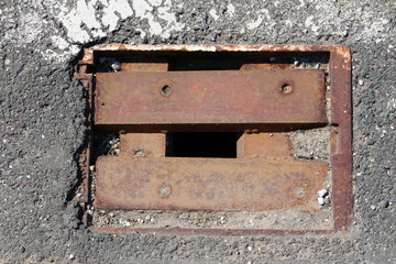 Square rusted iron drain opening with missing grates cover surrounded with old cracked asphalt on...