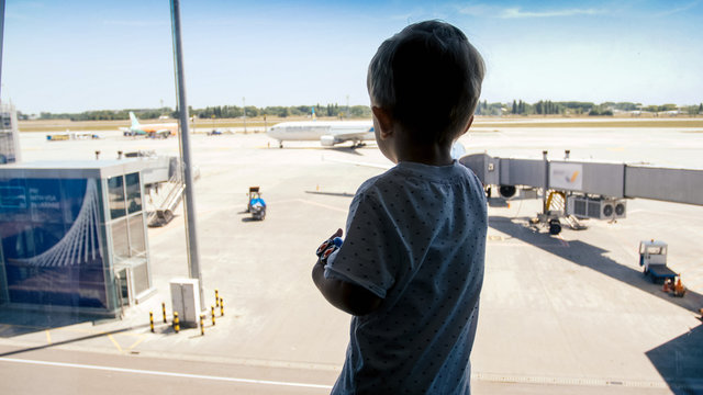 Silhouette of little toddler boy looking on airplanes through big window in airport