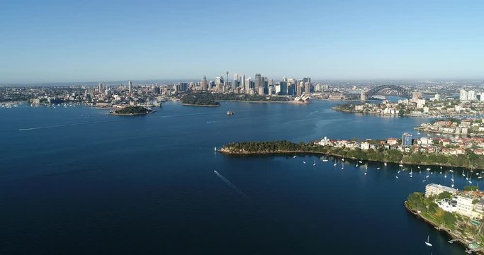 Cityscape of Sydney on harbour shores from distant city CBD landmarks aerial flying backward towards Mosman suburb.
