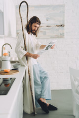 focused Jesus with wooden staff reading business newspaper in kitchen at home