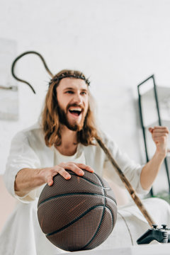 Excited Jesus With Wooden Staff Gesturing By Hand And Watching Basketball Match On Sofa At Home