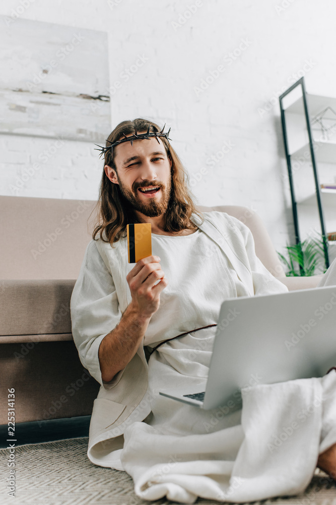 Canvas Prints cheerful Jesus showing credit card, winking and doing online shopping on laptop near sofa at home