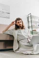 confident Jesus in crown of thorns sitting on floor and using laptop near sofa at home