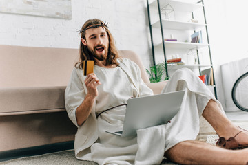 happy Jesus showing credit card, winking and doing online shopping on laptop near sofa at home