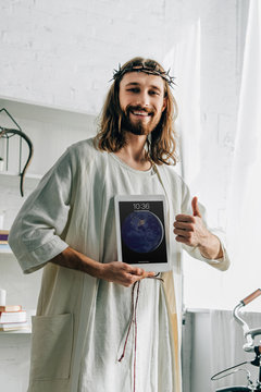 smiling Jesus in crown of thorns doing thumb gesture and showing ipad tablet at home