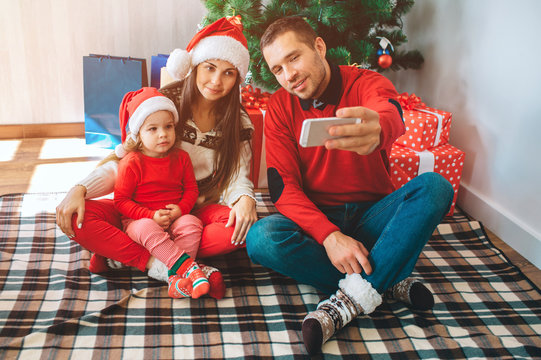 Merry Christmas and Happy New Year. Young man sits besides woman and child. He holds phone and takes selfie. Woman and kid lok at it and pose. Family wears Christmas clothes and hats.