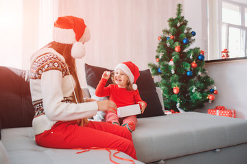 Merry Christmas and Happy New Year. Picture of happy kid sitting on sofa and hold white small round sweet. She looks at mother and smiling. Woman with long hair look at child. She is careful.