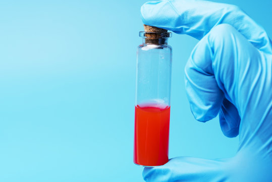 A gloved hand holds a test tube of red liquid on a blue background.