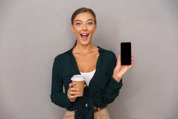 Business woman posing isolated over grey wall background drinking coffee showing display of mobile phone.