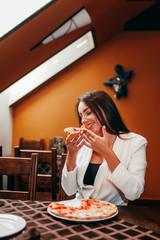 Beautiful stylish brunette girl in white jacket eating pizza at restaurant.