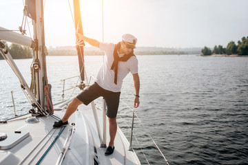Beautiful icture of handsome and serious sailor standing on his yacht. He holds on tube and look at edge of boat. Young man poses. Sun goes down. - obrazy, fototapety, plakaty