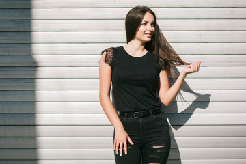 Fototapeta premium A brunette female with long hair wearing a blank black t-shirt i while standing on a concrete wall background on a street. Empty space for text or design.